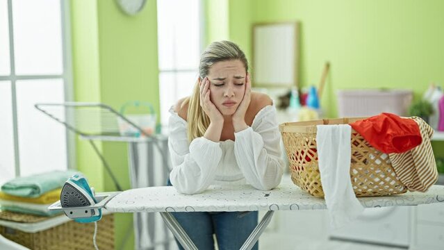 Young blonde woman leaning on ironing table with sad expression at laundry room