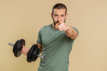 I am watching you. Athlete sportive man pointing at eyes and camera, show I am watching you gesture, spying on someone start working out. Middle eastern sportsman guy with dumbbell on beige background
