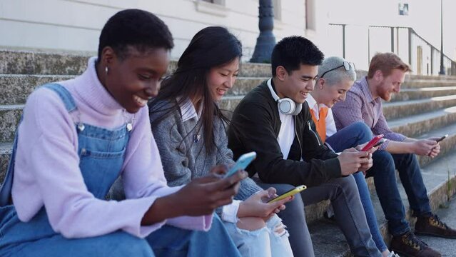Young group of people using mobile phone together sitting outdoors. Millennial diverse college students having fun watching social media content on smartphone app at college campus.