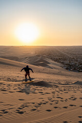 Backlit girl sandboarding as the sun sets over a city in Peru surrounded by sand dunes seen below.