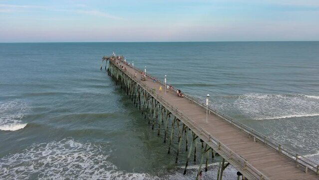 orbit aerial footage of Kure Pier with people fishing, ocean water, waves and condominiums and homes along the coast with blue sky and clouds at sunset in Wilmington North Carolina USA