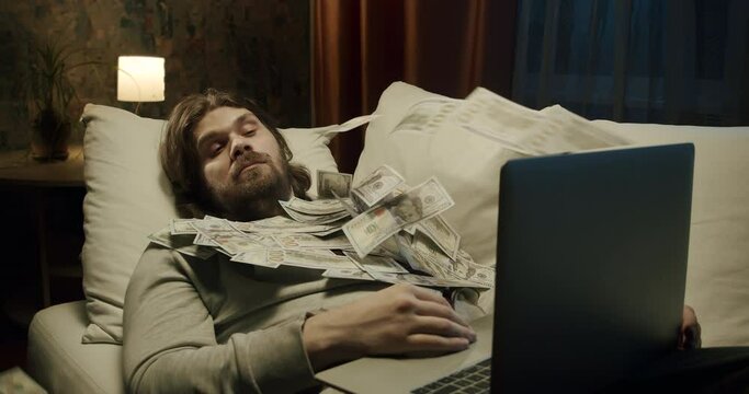 A man making money online while he sleeps. A man is awakened from sleep by a flow of money flying from his laptop. Earning money in sleep concept. Lazy man making money online lying on the couch. Cash