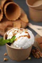Tasty ice cream with caramel sauce, mint and nuts in paper cup on grey wooden table