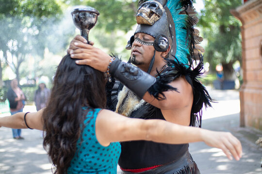 Aztec shaman making a healing ritual to an unrecognizable woman, using a chalice or calyx; traditional mayan religion dancer using a plume headdress with feathers