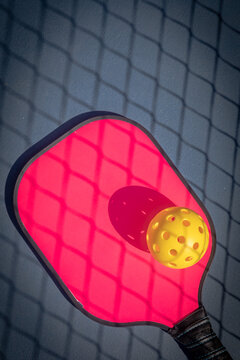 Pink Pickleball paddle with yellow Pickleball with shadow of Pickleball net.