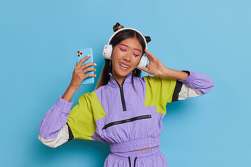 Asian student girl wearing stylish sport suit listening to music with white headphones on blue studio background, copy space, nice time concept