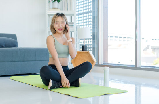Asian young female athlete teenager in sportswear sport bra sitting smiling on yoga pilates mat holding using smartphone in hand in gym after training exercising working out.