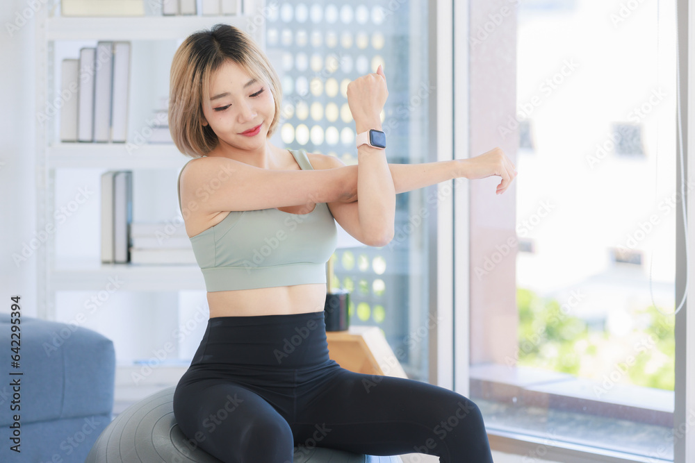 Wall mural Asian young sexy pretty fit female athlete teenager in sportswear sport bra leggings and watch sitting smiling stretching arms and hands on exercise ball warming up working out in living room at home