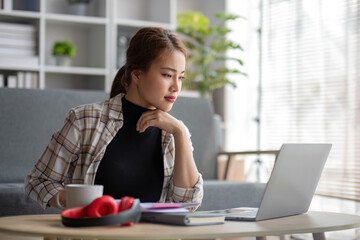 Beautiful and inspired Asian woman working on her tasks on laptop in a living room on the weekend.