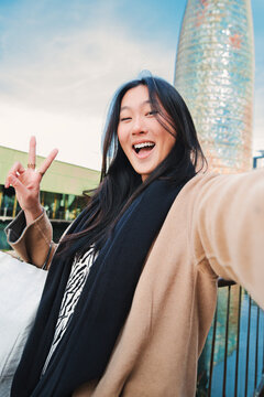 Vertical individual close up portrait of a happy asian young lady taking a selfie photo with an smartphone. Self portrait of chinese smiling woman doing the peace sign after shopping outdoors. High