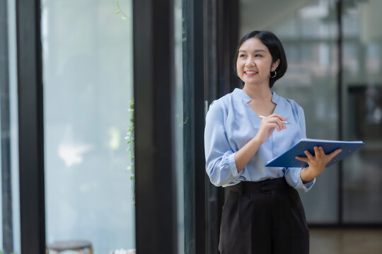 Happy Asian woman holding clipboard at office. Attractive and cute Asian businesswoman thinking up new ideas and holding clipboard at office.