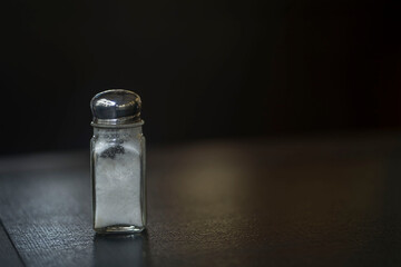 A close-up photo of a glass bottle filled with salt on a brown wooden table with a bokeh background.