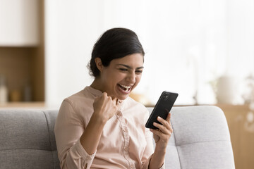 Successful excited young Indian woman shouting for joy at mobile phone, laughing, making yes fist,...