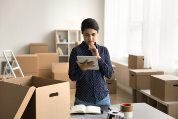 Serious Internet store Indian employee girl packing parcel for customer, using work application on tablet computer, holding gadget, standing at open box for transportation, shipping service