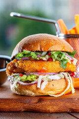 Hamburger with chicken cutlet, fresh tomatoes, red onion, lettuce and tomato sauce. Next to the burger is a metal mold with fries, a metal mold with chili sauce. Burger, fries and sauce are on a brown