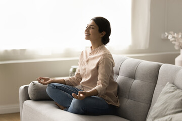 Happy peaceful young Indian yogi woman meditating on home couch, keeping lotus yoga pose, zen...