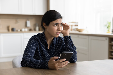 Concerned young Indian woman holding smartphone, looking away in deep thoughts, thinking on problems, feeling doubt, uncertain, worried, anxious, touching head, chatting at home table
