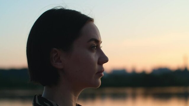 Beautiful woman face profile. Close-up view of pretty dreamy girl looking out at sunset in front of the river, nature landscape view. Slow motion 4K