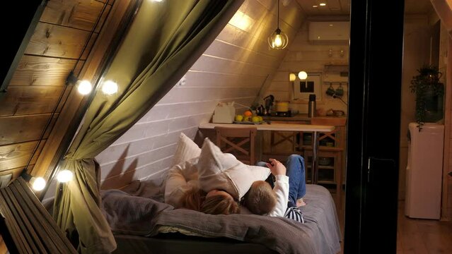 Mother and son have fun lying on the bed in a small wooden triangular house, they throw pillows up. Vacation in a country house in autumn. View through a window with flashlights and a thick curtain.