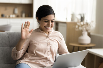 Happy friendly Indian freelancer woman waving hand hello at monitor, saying hi with greeting gesture, talking on video call, smiling, using laptop computer for Internet communication