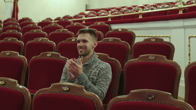 European man in sweater sits alone in stalls in theater auditorium. Theatergoer smiles and applauds, waiting for start of the performance in opera house with luxury interior.