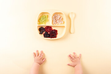 Children's silicone plate with food, the baby's hands reach for dinner. Feeding on a yellow background.