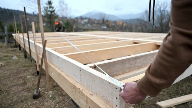 Man worker building wooden frame house on pile foundation. Close up carpenter using tape measure for measuring wooden planks. Carpentry concept.
