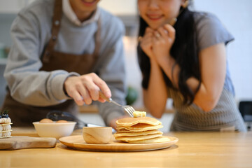 Happy young couple man preparing breakfast for his girlfriend in cozy kitchen, putting butter on the hot pancakes