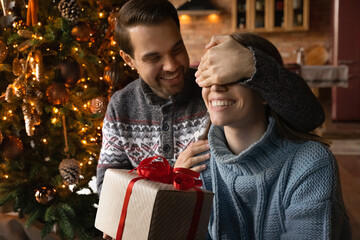 Smiling young man making surprise to beautiful wife, presenting christmas gift in wrapped box,...