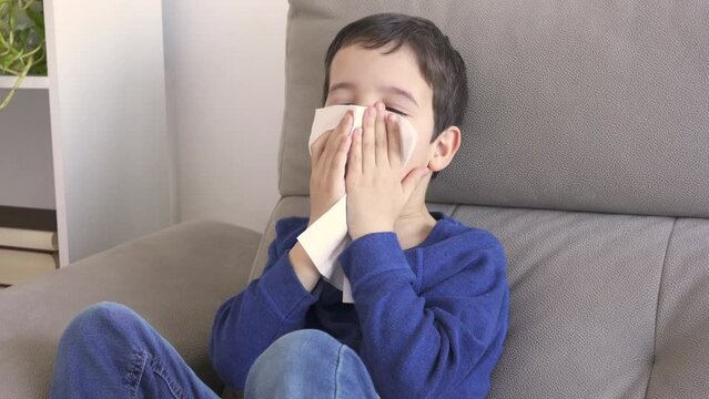 Child blowing in a wipe suffering flu symptoms sitting on a sofa at home in winter