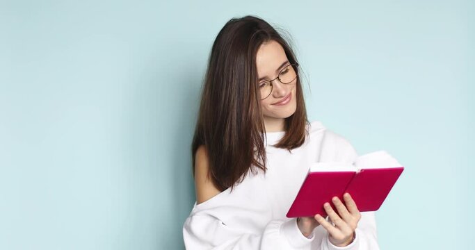 Young beautiful girl read in pink diary her romantic story, poem while she is smiling, dreaming, thinking. Woman in eyeglasses remember some memory, while read in her pink notebook on blue background.