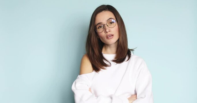 So what. Young caucasian woman in eyeglasses crossed her arms, looking at camera with doubt and shaking palm, waiting for explanations, isolated on blue studio background.
