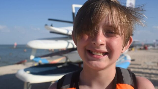 smiling child  in a life jacket at the surf station on the beach in summer. joyful 10 year old child stands next to the windsurf boards, waiting for a workout.