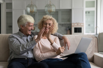Cheerful satisfied elder husband and wife giving high five at laptop with pride, celebrating...