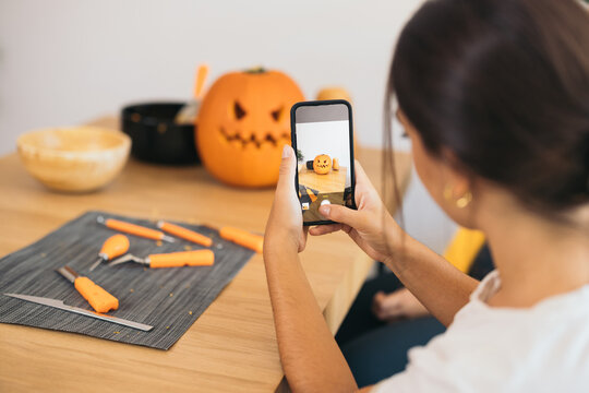Crop young ethnic woman taking picture of Halloween pumpkin with smartphone