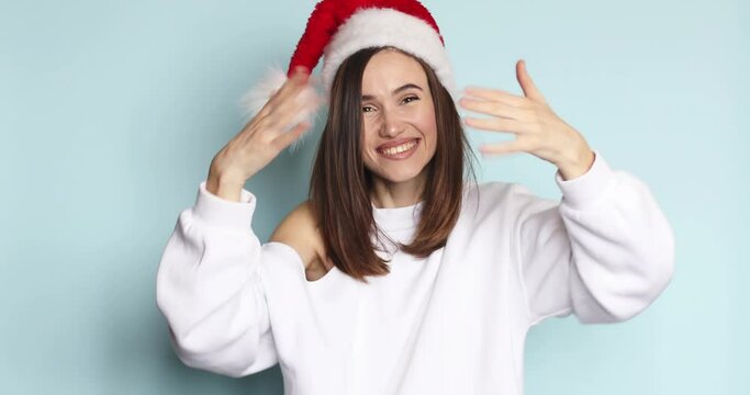 Come here, follow me, quickly! Beautiful young brunette woman in Santa hat, waving hand, making beckoning gesture with hands, inviting to approach, looking playful flirting on blue background.