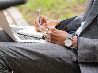 Unrecognizable businessman writing in notepad with pen while sitting with laptop in daylight