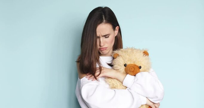 Depressed frustrated young woman standing with teddy bear looking at camera with sad facial expression. Portrait of upset beautiful slim caucasian lady posing at blue background hugging toy.