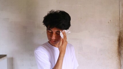 an asian man in a white t-shirt is wiping his face with a tissue