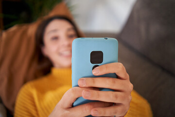 Close-up unrecognizable girl hands using phone lying on living room sofa. Unfocused woman looking at mobile at home. Focus on cell in blue case. Young people addicted to technological devices. - obrazy, fototapety, plakaty