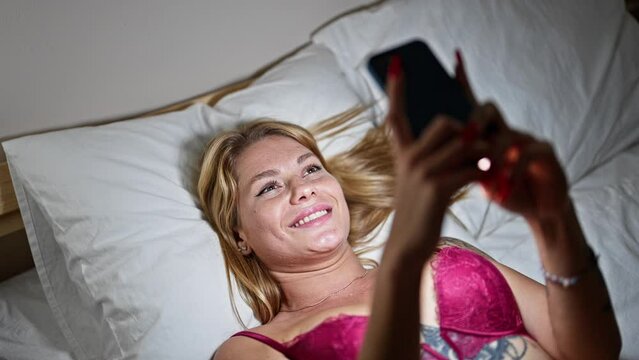 Young blonde woman wearing sensual lingerie lying on bed using smartphone at bedroom