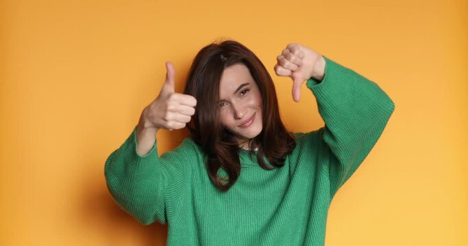 Young woman in green sweater making good-bad sign isolated on yellow background. Girl make choice, or makes a decision thumb up or thumb down, like or dislike, yes or no. 