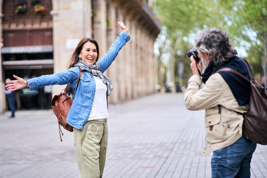 Excited mature woman posing funny for photo taken with professional camera by gray man on city street. Couple cheerful tourists enjoying their retirement holiday. Love and romantic getaways in old age