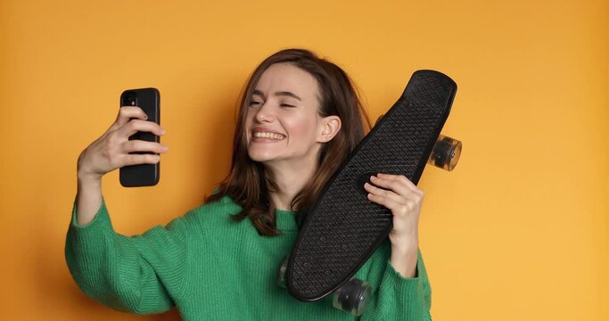 Brunette girl stares at the phone and how penny board, make selfie. Female uses phone like mirror. Young girl make photo from hands with phone, show tongue, posing on yellow background.