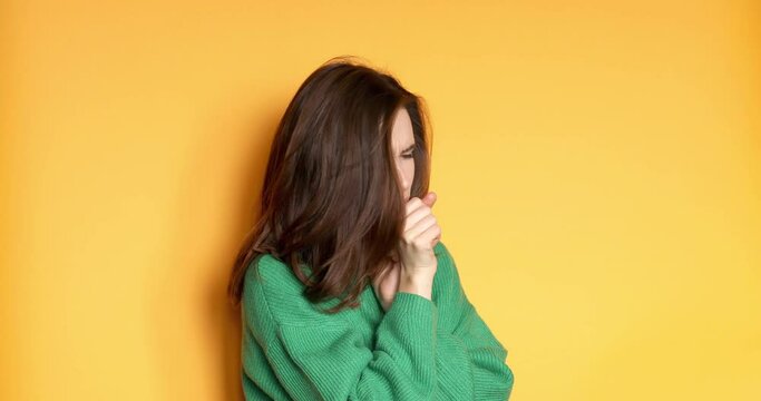 Woman wear green sweater is suffering with cough and feeling bad on yellow background. Health care and safety. Millennial woman coughing hardly while hold neck.