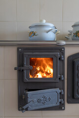 Wood burning tiled stove with fire in the kitchen with white pots on top.