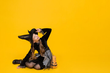 A little witch in a pointy hat sits on the floor and looks at your advertisement. A magical girl in a black carnival dress. Happy Halloween. Yellow isolated background. Copy space. Banner.
