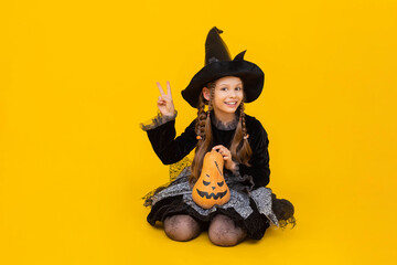 A little girl in a witch costume and a pointed hat is sitting in full height, legs tucked under her, and holding a pumpkin in her hands. Child is happy with Halloween. Yellow isolated background.