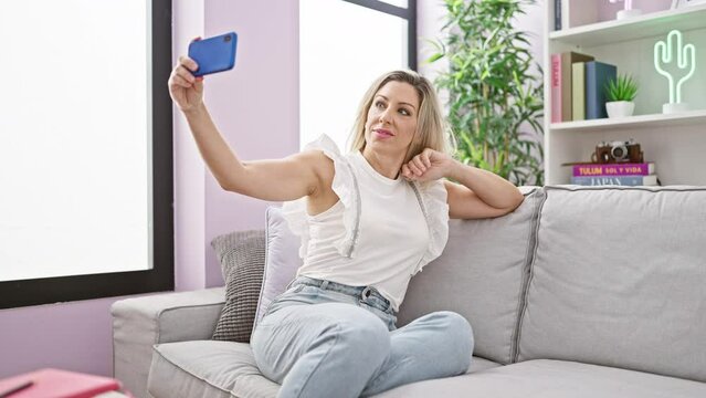 Young blonde woman taking selfie picture with smartphone sitting on the sofa at home