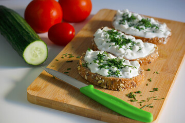 Close-up of three slices of wholemeal bread spread with butter and cottage cheese sprinkled with freshly chopped chives on a bamboo cutting board, tomatoes and cucumber in the background. Healthy food
